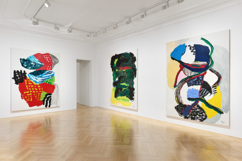 An art gallery displays three large, colorful abstract paintings on white walls. The room features wooden parquet flooring and ceiling track lighting.
