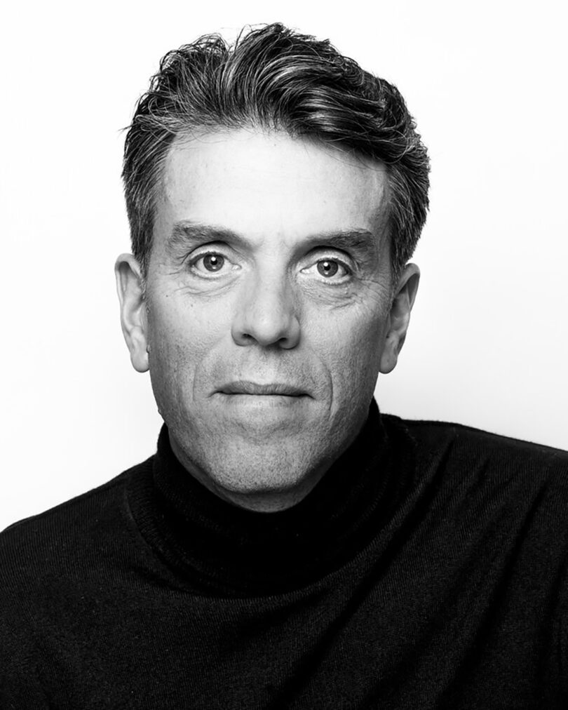 Black and white portrait of a man with short, dark hair, wearing a black turtleneck, looking directly at the camera against a plain white background.