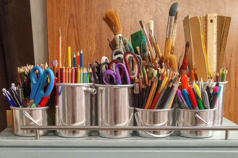 A collection of scissors, colored pencils, paintbrushes, and other art supplies organized in metal containers on a shelf.