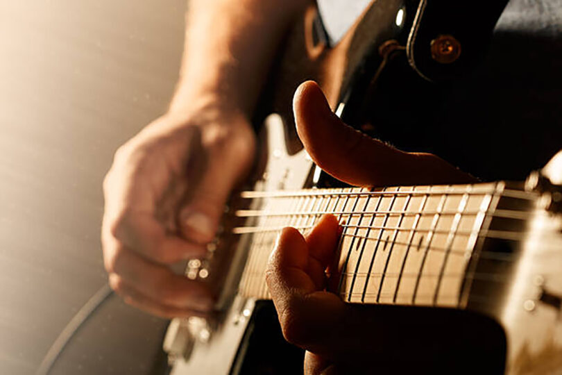 Close-up of a person playing a black electric guitar, focusing on their hands and fingers pressing the strings. The background is softly lit.