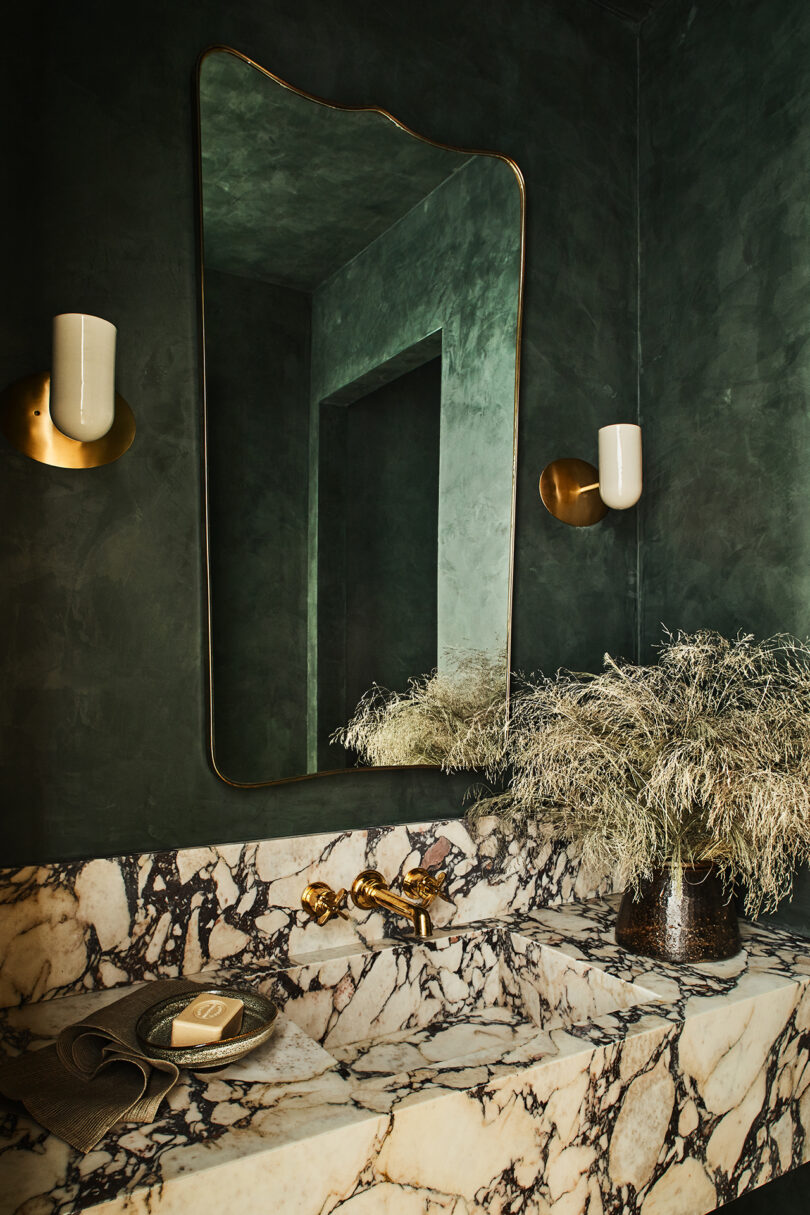 A marble sink with gold fixtures sits below a large mirror and two wall-mounted lights in a dark green bathroom with a small plant on the countertop.