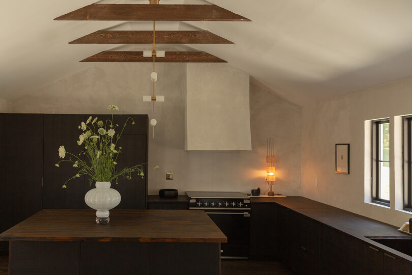 A minimalist kitchen with dark wood countertops, a white vase with flowers on an island, a black oven with a range hood, and pendant lights hanging from an exposed beam ceiling.