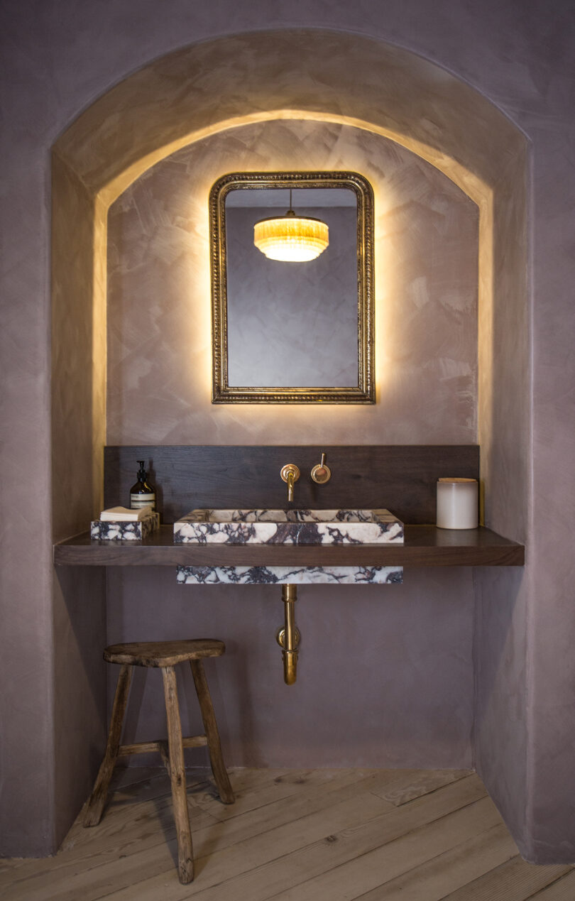 A bathroom with a marble sink, wooden stool, framed mirror, and a lit ceiling light. The walls and ceiling have a textured taupe finish.