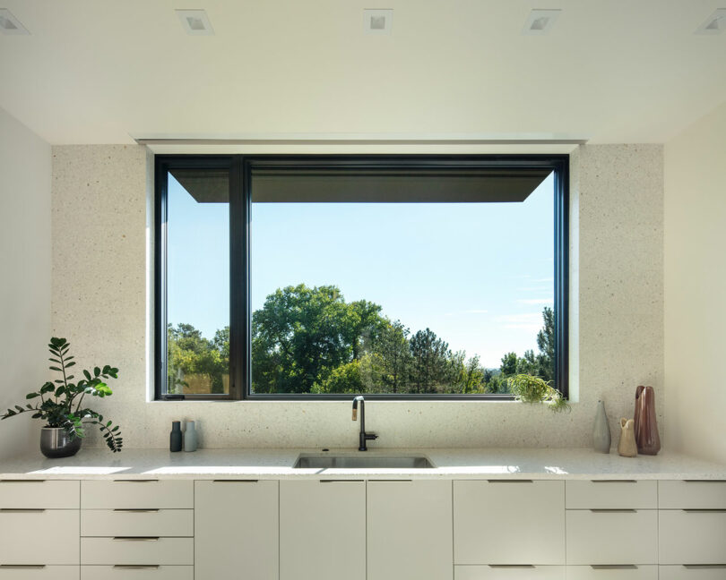 A modern kitchen with a large window showcasing an outdoor view. The countertop features a black faucet, a plant, a ceramic vase, and various small containers.