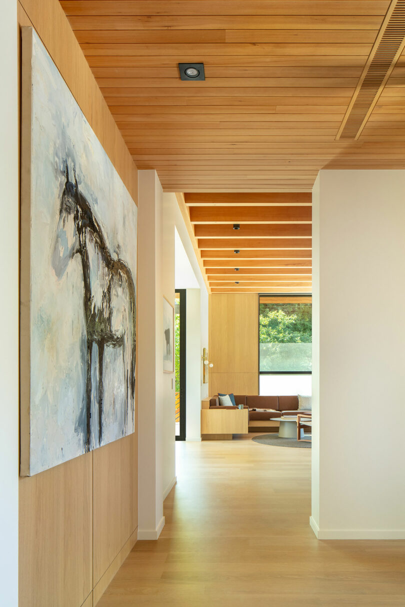 A modern hallway with wooden flooring and ceiling, a painting of a horse on the wall, and a view into a living area with natural light streaming through large windows.