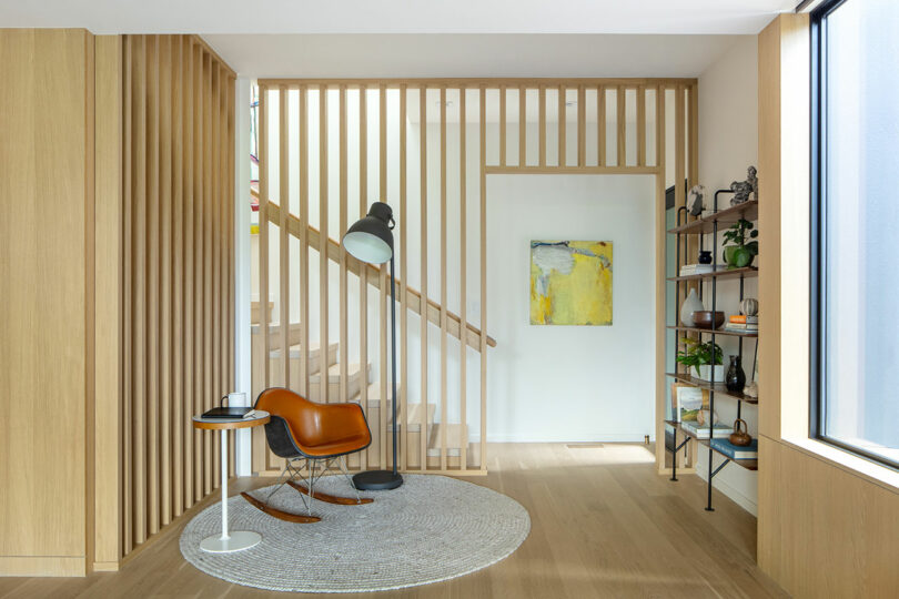 A modern, minimalist living space with a wooden aesthetic features a rocking chair, a tall floor lamp, a circular rug, wall art, and a staircase behind vertical wooden slats.