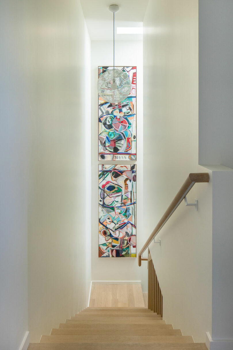 A staircase with wooden steps and a handrail leads down to a colorful abstract painting on the wall. A pendant light hangs from the white ceiling, illuminating the space.