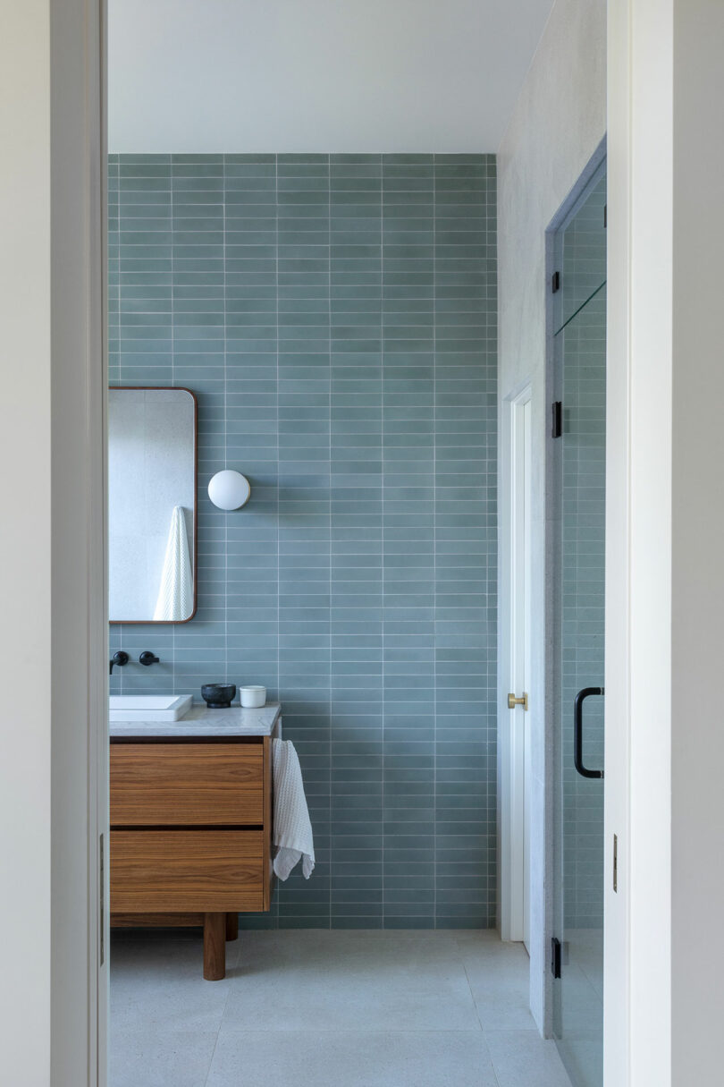 A bathroom featuring a wooden vanity with a white sink, a rectangular mirror above it, light blue vertical tiles on the wall, and a glass shower door to the right.