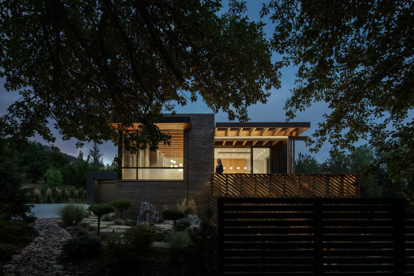 A modern house with large windows and a wooden deck is seen at dusk, surrounded by trees and landscaped garden, with warm interior lights glowing inside.