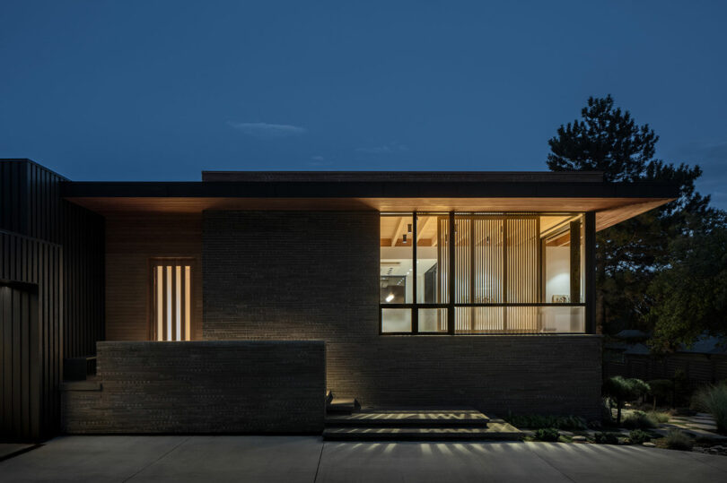 Modern house with large windows illuminated from within at dusk. The structure has a flat roof, brick walls, and a minimalist design with surrounding greenery.
