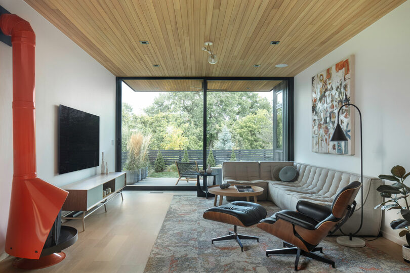 Modern living room with wooden ceiling, large glass doors leading to an outdoor area, gray sectional sofa, Eames lounge chair, abstract artwork, and orange hanging fireplace.