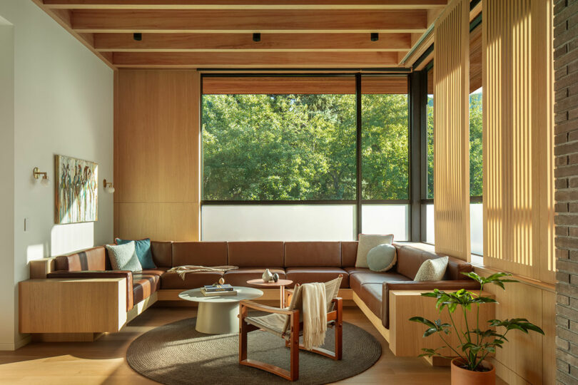 A modern living room with wood accents, a large sectional sofa, a round coffee table, a wooden armchair, a painting, and large windows offering a view of greenery.