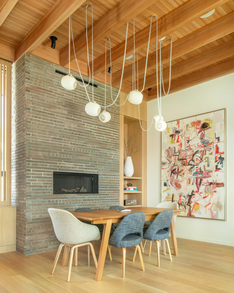 A dining area with a wooden table, four chairs, modern light fixture, brick fireplace, abstract painting, and wooden ceiling beams. Decor elements include books and a vase with branches.