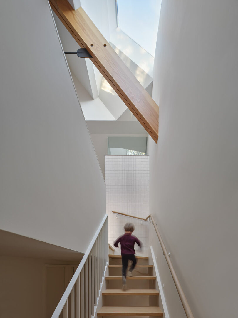 A person in a red shirt runs up a white staircase inside a modern building with a wooden beam and a skylight above.