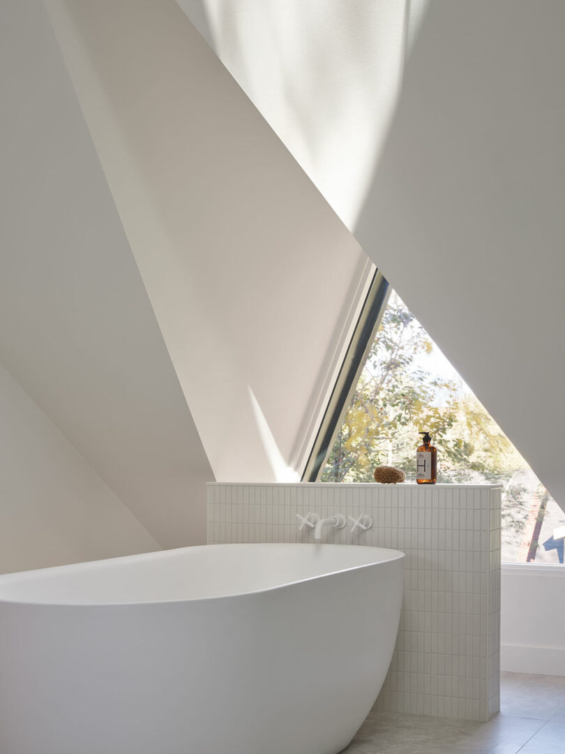 Modern bathroom with a white freestanding bathtub, geometric walls, a large window, and natural light streaming in. The tub's edge holds a bottle and a sponge.