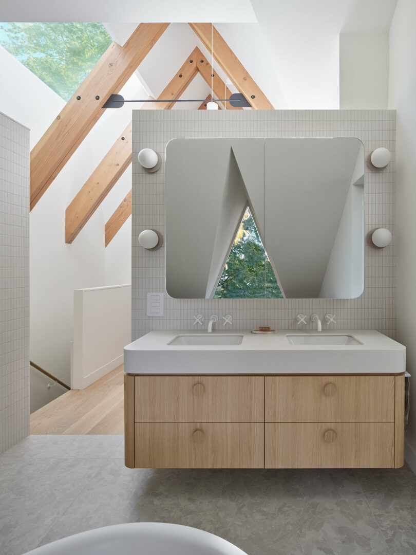 A modern bathroom featuring a double-sink vanity with light wood cabinets, large mirror, and a geometric window. The staircase and wooden beams are visible in the background.
