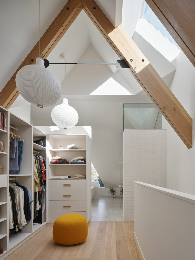 A modern, minimalist walk-in closet with open shelving, a yellow ottoman, and slanted wooden ceiling beams. Natural light enters through skylights, illuminating the space.