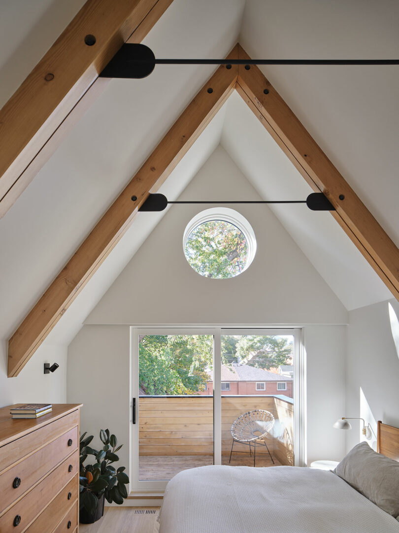 A bedroom with a vaulted ceiling features wooden beams, a circular window, sliding glass doors, a chest of drawers, a bed, and a balcony with an outdoor chair.