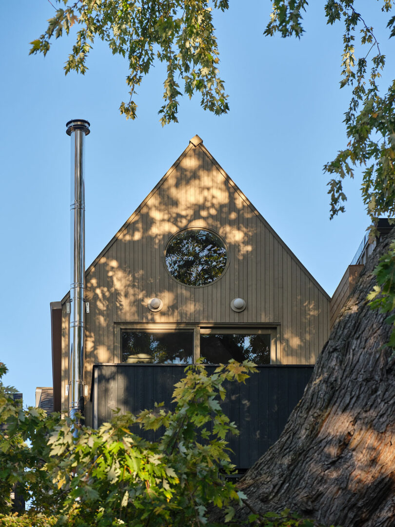 A modern, triangular wooden house with a circular window and two smaller square windows. A metal chimney is visible on the left side. Tree branches frame the image against a clear blue sky.