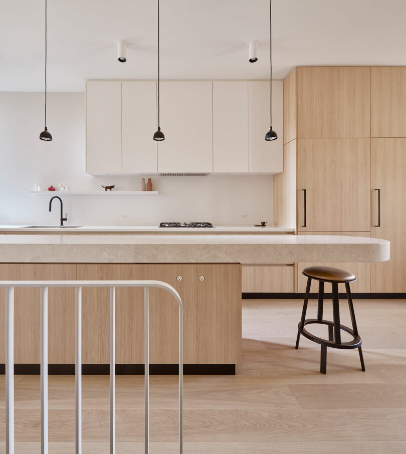 A modern kitchen features light wood cabinetry, a white island with a countertop, hanging pendant lights, a black stool, and minimalistic decor.