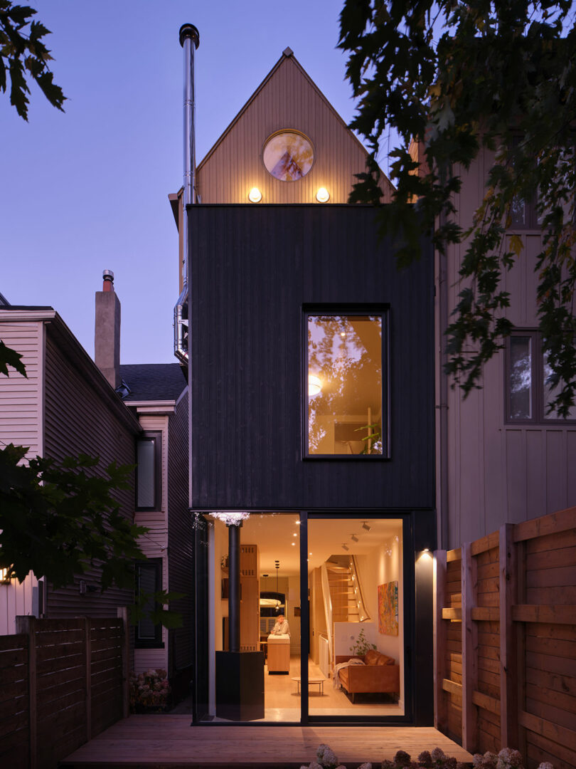 A three-story modern house with a dark exterior and large windows, illuminated at dusk. The bottom floor features sliding glass doors leading to a well-lit interior with visible stairs.