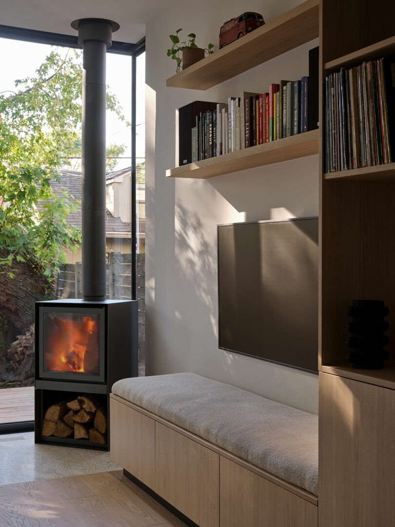 A modern living room with a cozy bench, a wood-burning stove, shelves with books and decor, and a wall-mounted TV. Large windows reveal a tree-filled outdoors. Wood logs are stored below the stove.