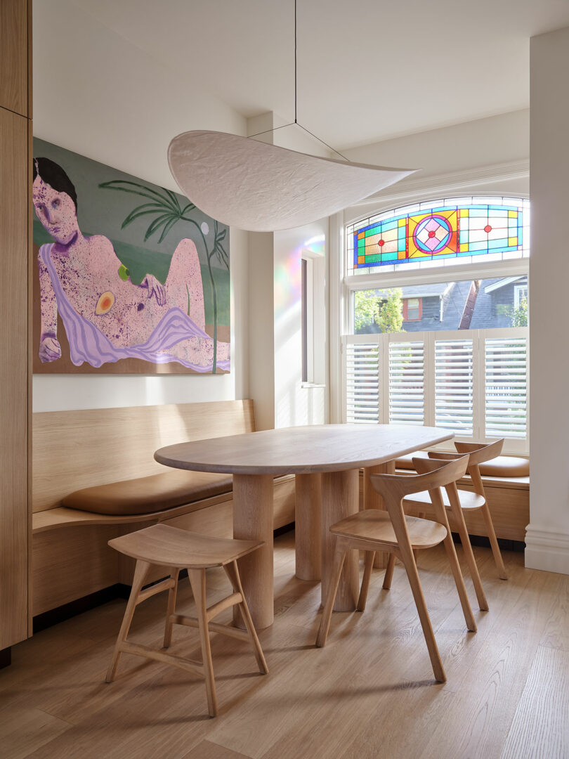 A modern dining area features a wooden table with four chairs, a cushioned bench, a large abstract painting, a sculptural ceiling light, and a window with colorful stained-glass details.
