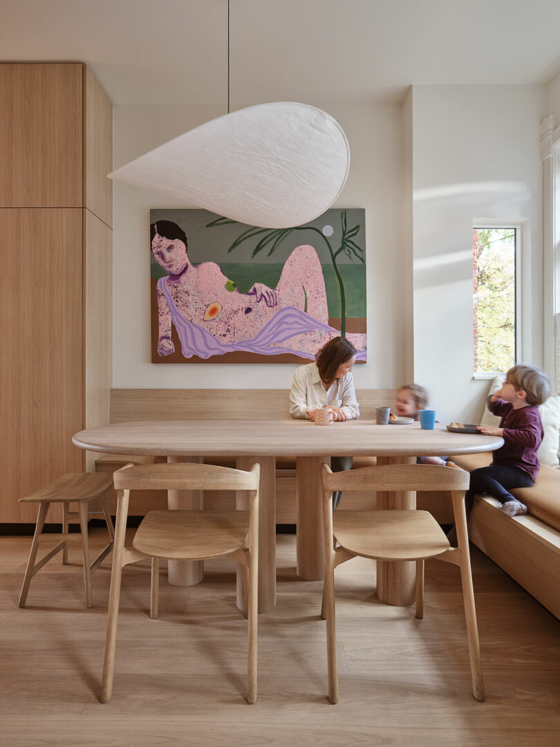 A person sits at a wooden dining table with two children on a bench in a modern, minimalist dining room. A large painting of a reclining figure hangs on the wall behind them.