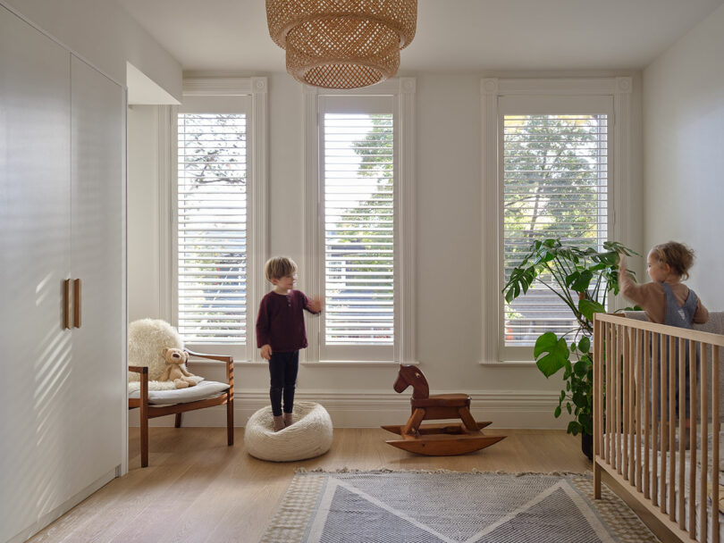 A child stands on a chair and another by a crib in a bright room with three tall windows, a wooden rocking horse, a large green plant, a beige rug, and a wicker ceiling light fixture.