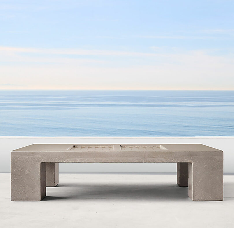 A minimalist concrete bench designed by James de Wulf with a rectangular shape is set on a polished concrete surface. The background features a clear, blue sky and a calm ocean.
