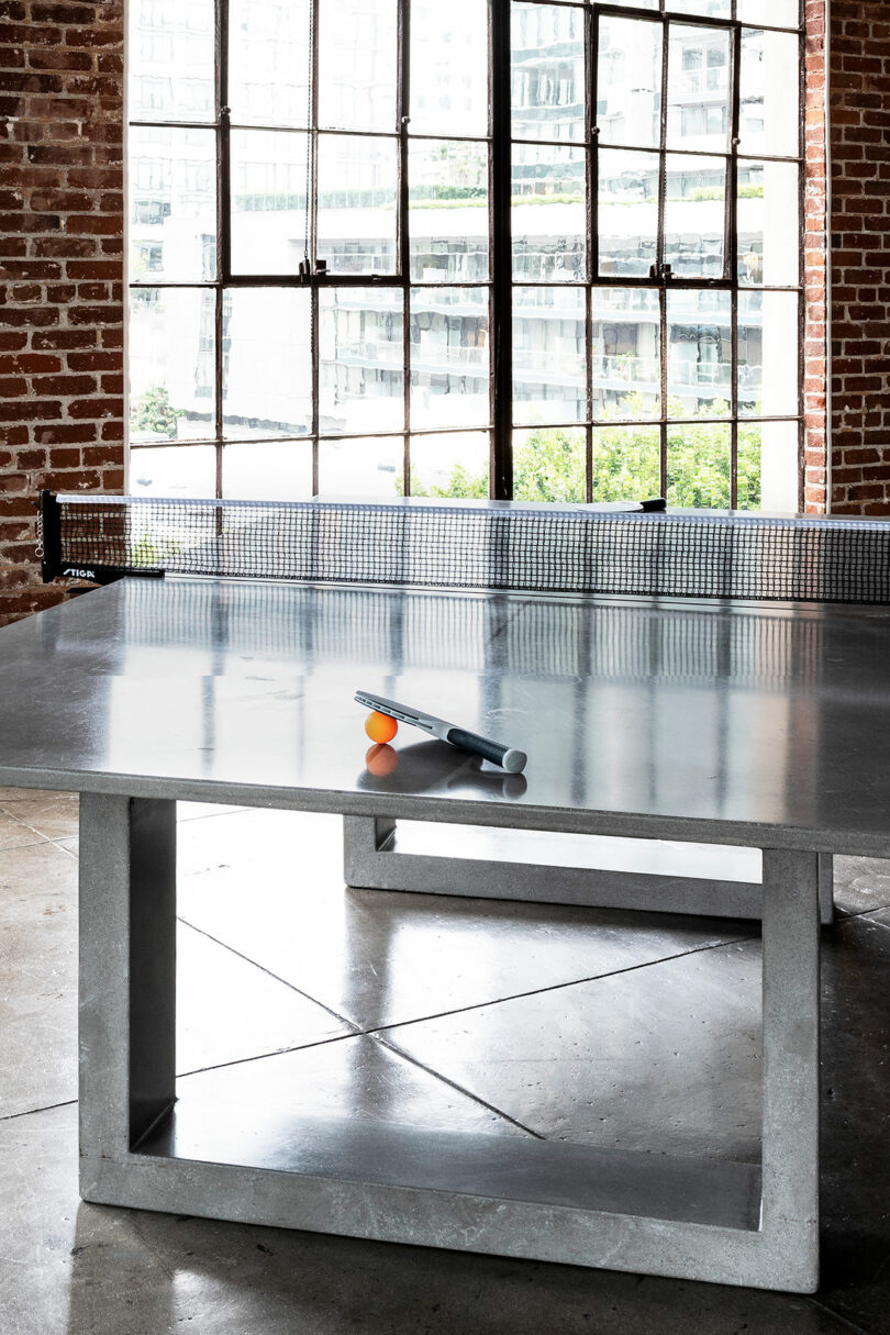 A modern James de Wulf ping pong table with two paddles and a ball on top, placed in a room with brick walls and large windows.