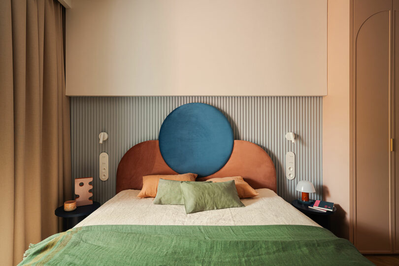 A modern bedroom with a colorful headboard featuring a blue circle and orange shapes. The bed is adorned with green and beige pillows and a green blanket. Nightstands with lamps and books are on either side.