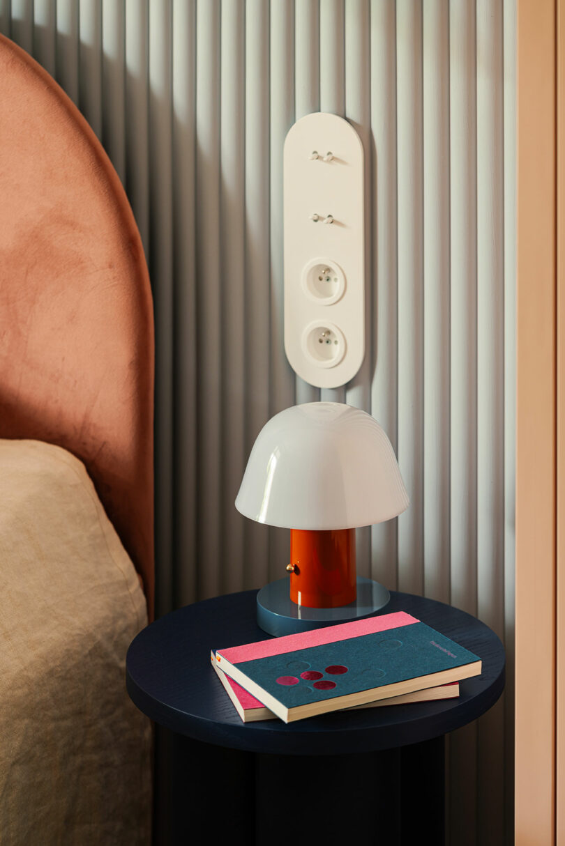 A bedside table with a white and orange lamp, two books with colorful covers, and a wall-mounted power outlet panel behind it. The bed with a beige blanket is partially visible.