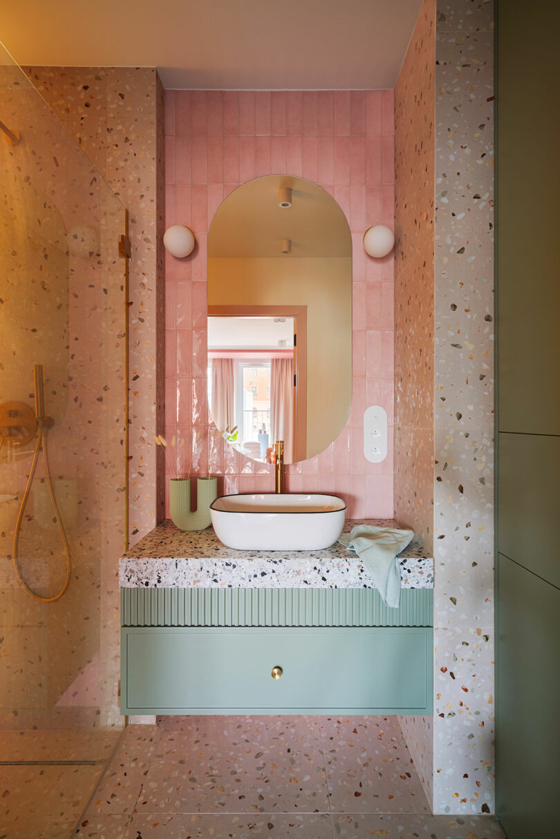 Modern bathroom with a pink and terrazzo design, featuring a circular mirror, a white sink, a green vanity cabinet with a gold knob, and a walk-in shower.