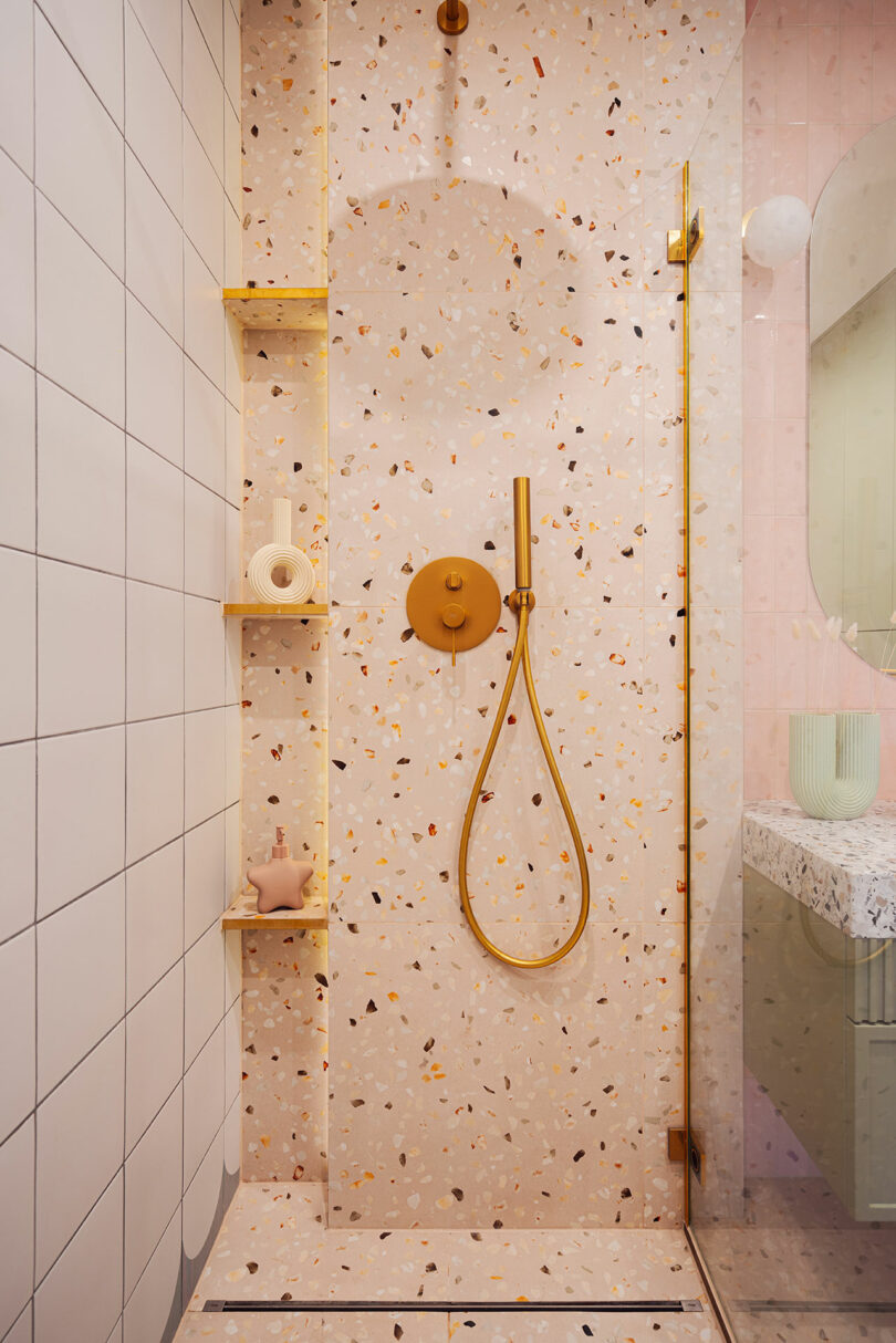 A modern shower with terrazzo walls, gold fixtures, an overhead and handheld showerhead, built-in corner shelves holding toiletries, and a glass partition.