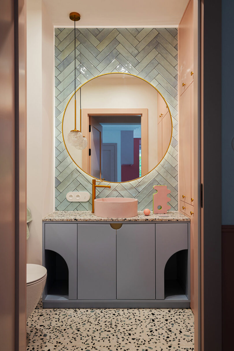 Modern bathroom with a large round mirror, pastel pink sink, blue cabinets, patterned floor tiles, and herringbone tiled wall. Warm lighting and minimal decor provide a clean, cohesive look.