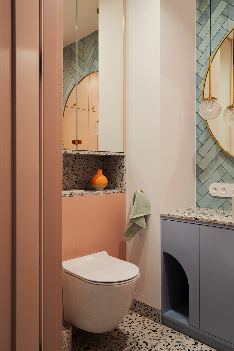 A modern bathroom with a wall-mounted toilet, pink and blue cabinetry, a terrazzo countertop and floor, a round mirror, and hanging lights. A folded green towel hangs on a silver hook beside the sink.