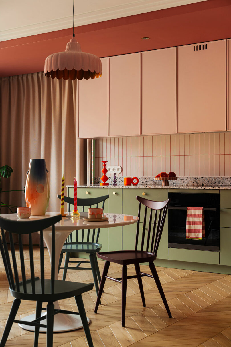 A modern kitchen with pink cabinets, green lower cabinets, and a white dining table with black chairs. A colorful vase and candles are on the table, and a striped towel hangs from the oven.