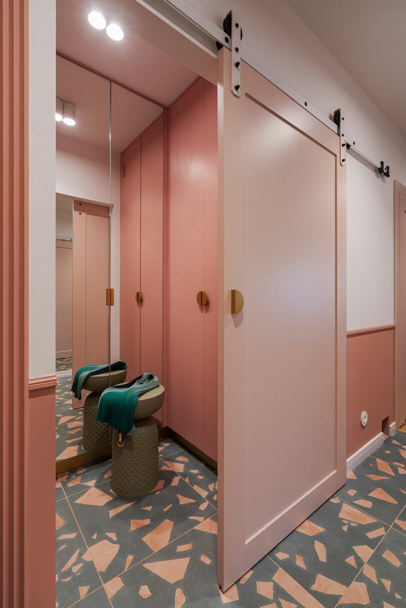 A modern hallway with pink sliding doors, geometric patterned floor tiles, and a small cushioned stool in front of a full-length mirror. Walls are painted in complementary pastel shades.