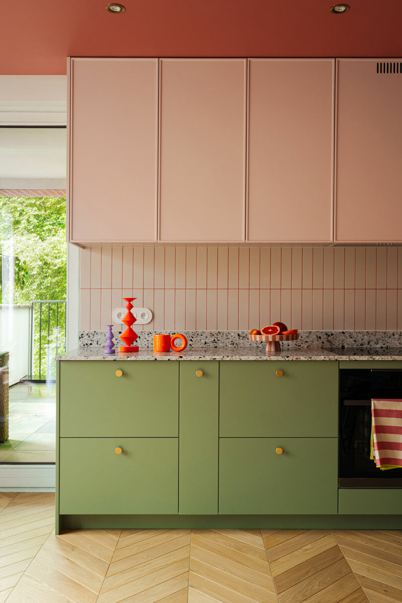 A modern kitchen with green lower cabinets, pink upper cabinets, and a terrazzo countertop. The backsplash tiles are pink. A few colorful items are on the counter. There is a patio door to the left.