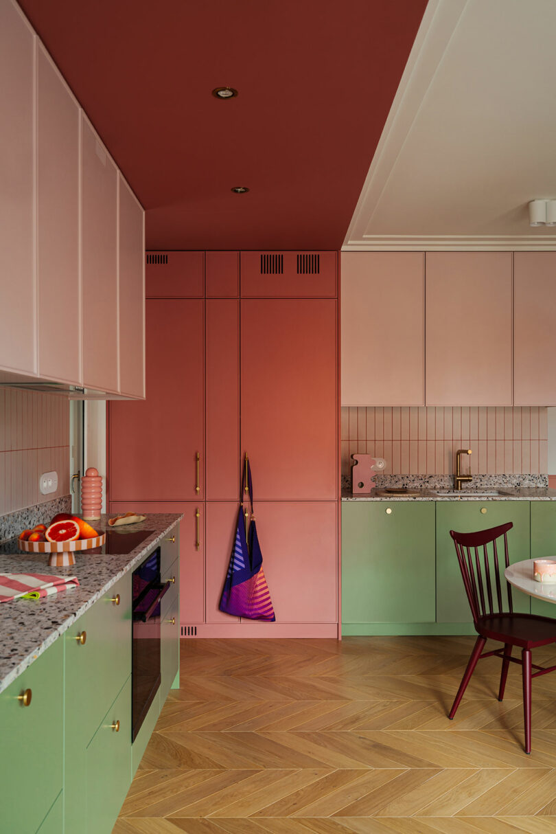 A modern kitchen with pink and green cabinets, marble countertops, a pink and green oven, wooden chevron-patterned floor, and a red ceiling. A multicolored apron hangs on a door.