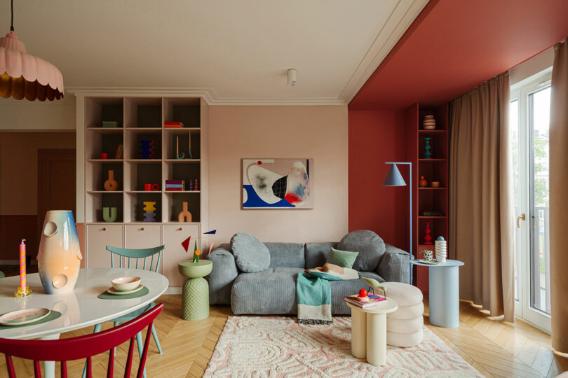 A modern living room with colorful decor, a grey couch, and built-in shelves. The room has a mix of red, pink, and neutral tones with various art pieces and a dining table with chairs.