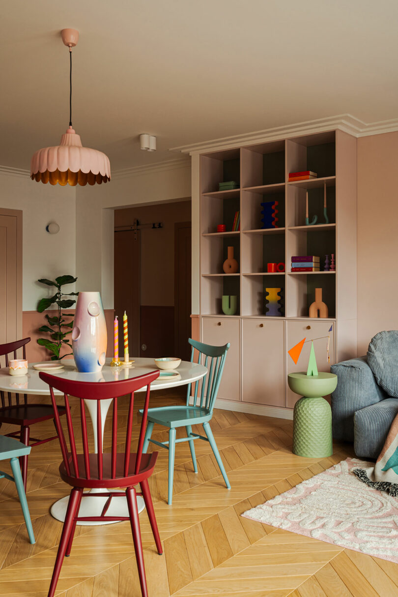 A colorful dining area with a round table, multicolored chairs, a pastel pink cabinet with shelves, geometric decor, and a potted plant in the background. The floor has a herringbone pattern.