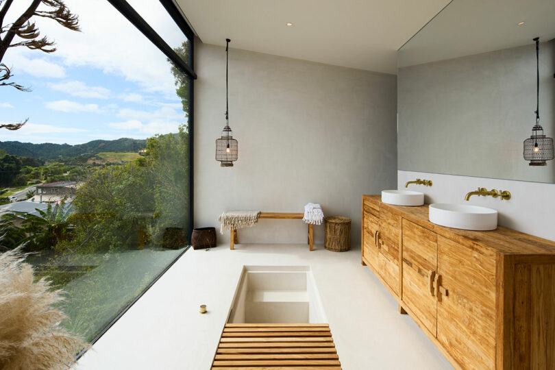 Modern bathroom with two vessel sinks on a wooden vanity, large mirror, pendant lights, a bench, and a recessed bathtub. Floor-to-ceiling windows reveal a view of greenery and hills.