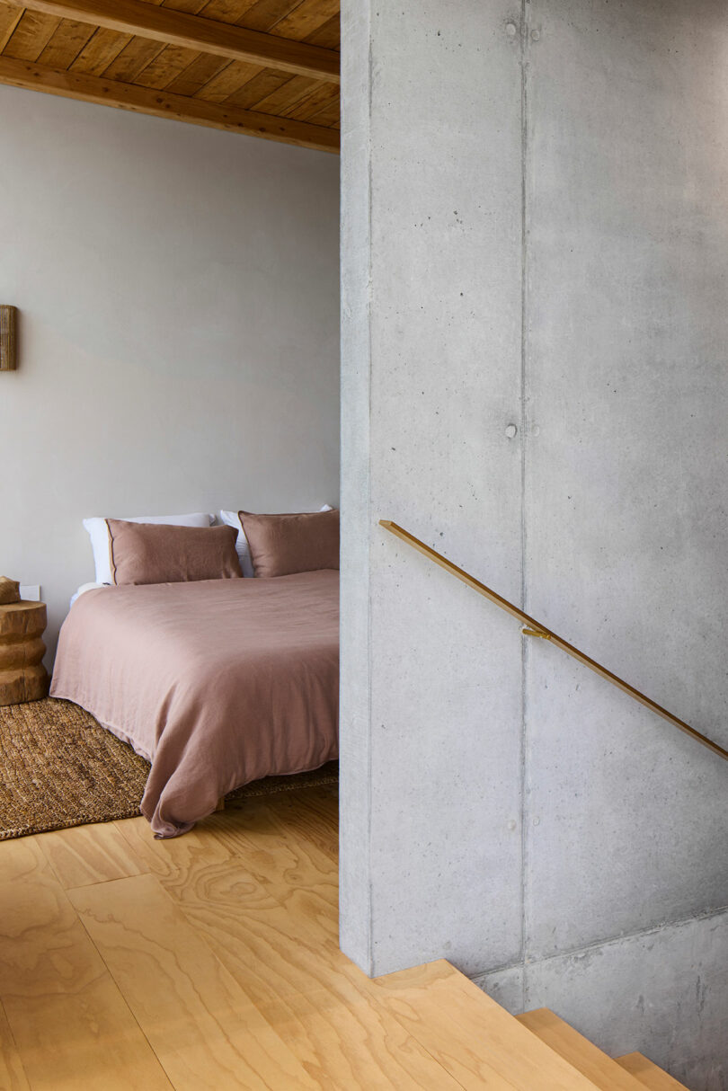 A bedroom with a wooden ceiling, a cozy bed with pink bedding, a woven rug, and a concrete wall with a wooden handrail leading down wooden stairs.