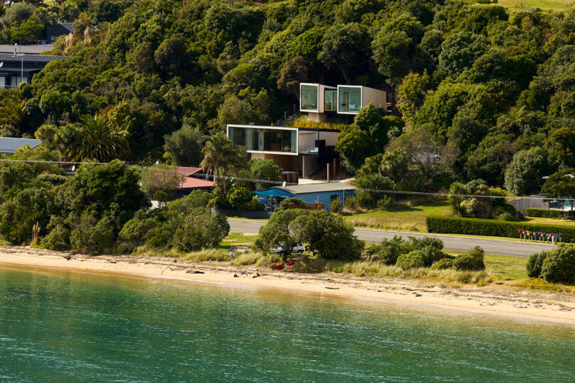 Modern hillside house with large windows and a multi-level design, surrounded by trees, overlooking a beach with calm turquoise waters and a nearby road.