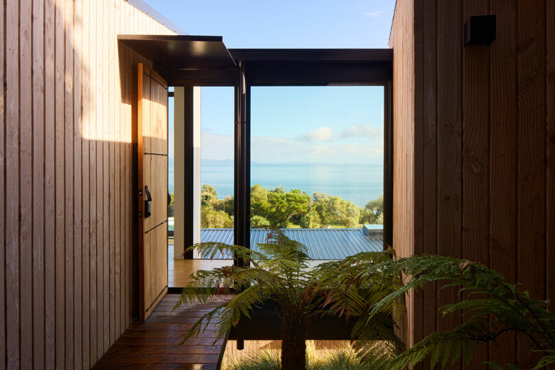 A wooden entryway with a large glass door opens to a scenic view of the ocean and trees, framed by interior wood paneling and lit by natural sunlight.