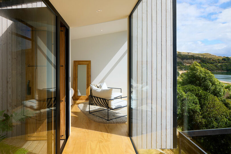A modern sunlit room with glass walls, featuring two chairs and a wooden floor, overlooks a scenic landscape with hills and a body of water.