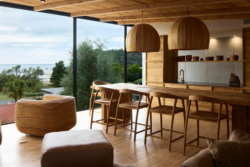 A modern, wooden-themed kitchen and dining area with large windows offering a view of trees and the ocean. The room features natural wood furniture and large, dome-shaped hanging lights.