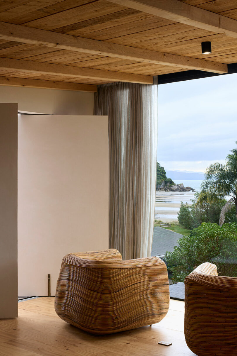 A modern wooden interior with large windows overlooks a scenic beach. Two unique, rounded wooden chairs are positioned near the entrance, adorned with flowing sheer curtains.